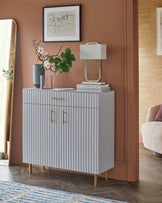 Stylish white cabinet with ribbed design, gold hardware, a table lamp, decorative vase, and a flower arrangement on top.