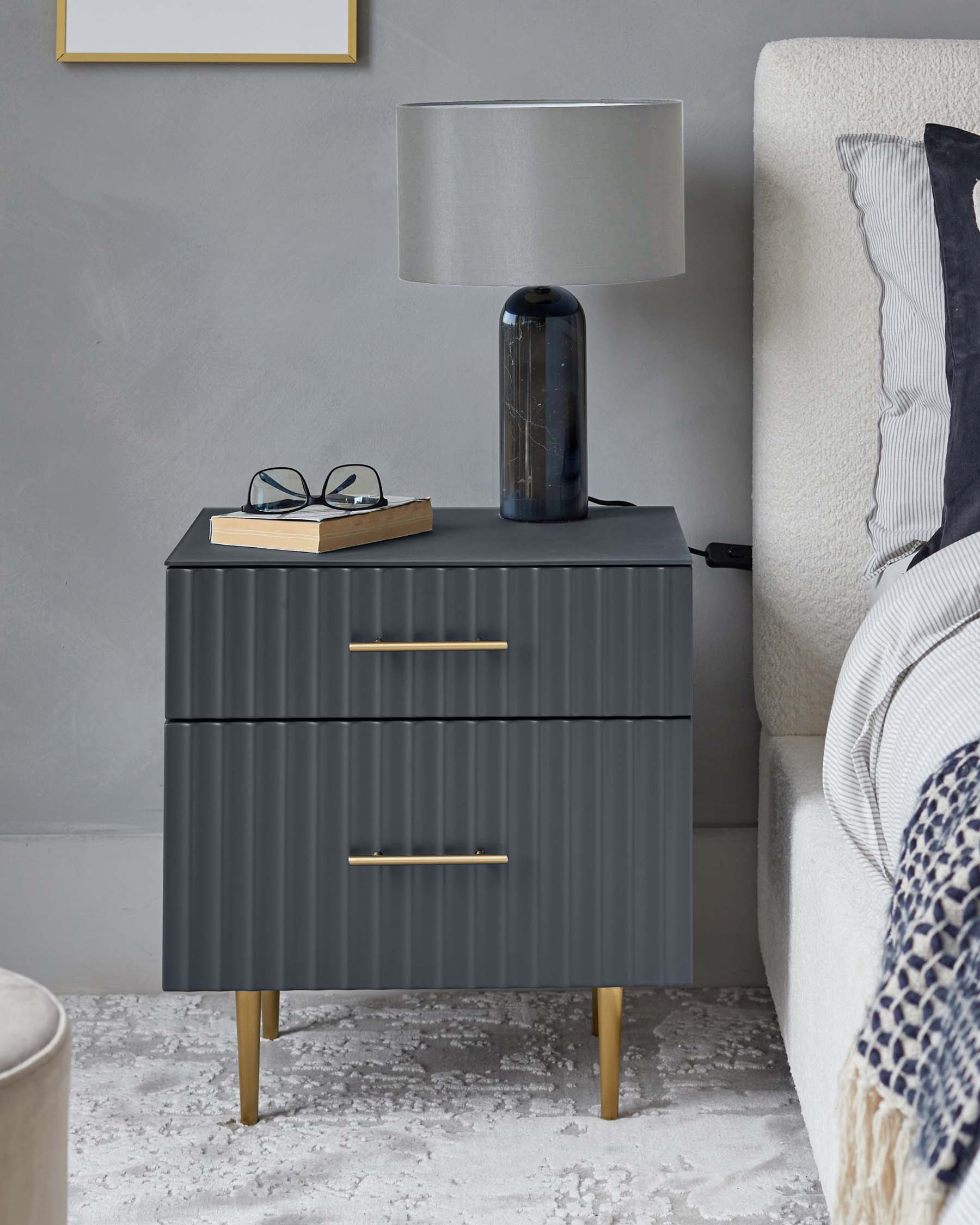 A modern dark gray nightstand with two drawers and gold handles, topped with a sleek lamp and a book.