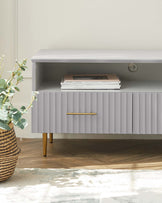 Modern minimalist style sideboard in grey with a smooth top surface and a ribbed front design. Features include a spacious open shelf with a collection of books and a metallic horizontal pull handle on the cabinet door. The sideboard is elevated by four tapered wooden legs with a golden finish, complementing the overall sleek appearance. The setting is further enhanced by a textured rug and a woven basket with a plant to the side, creating a cosy atmosphere.