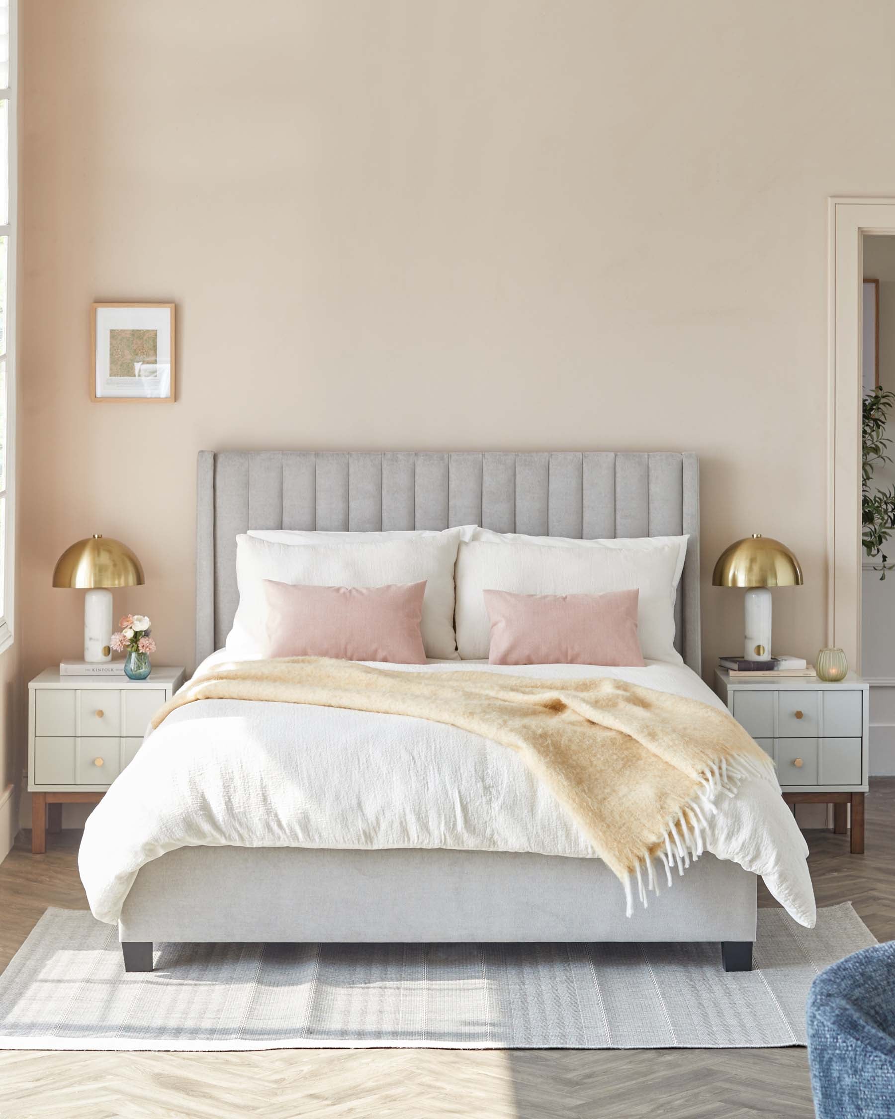 Elegant bedroom interior with a plush king-size bed featuring a tufted light grey headboard and a neutral-coloured bedding set. On either side, two modern white nightstands with gold knobs and matching gold dome table lamps complete the serene and sophisticated aesthetic. A soft caramel throw blanket adds a touch of warmth, while a simple grey area rug anchors the space. A framed artwork hangs on the wall, complementing the room's tranquil palette.