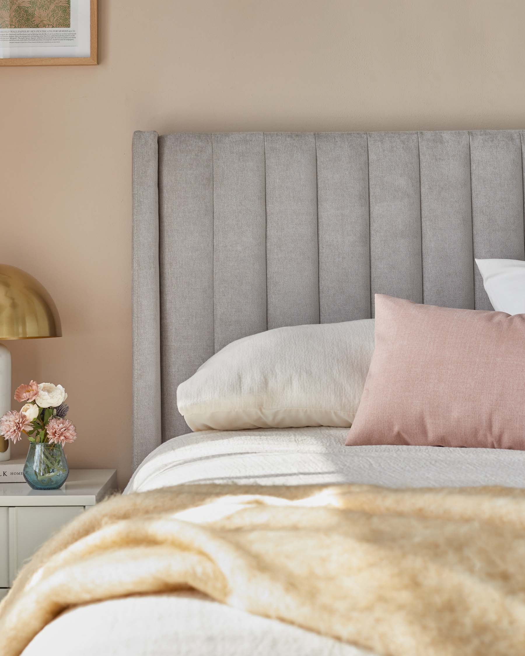 King-sized bed with a tall, channel-tufted headboard in a soft grey fabric. The bed is dressed with a white duvet, a tan throw blanket, and assorted pillows including white and soft pink colours. A small white nightstand is partially visible with a vase of pink flowers on top.