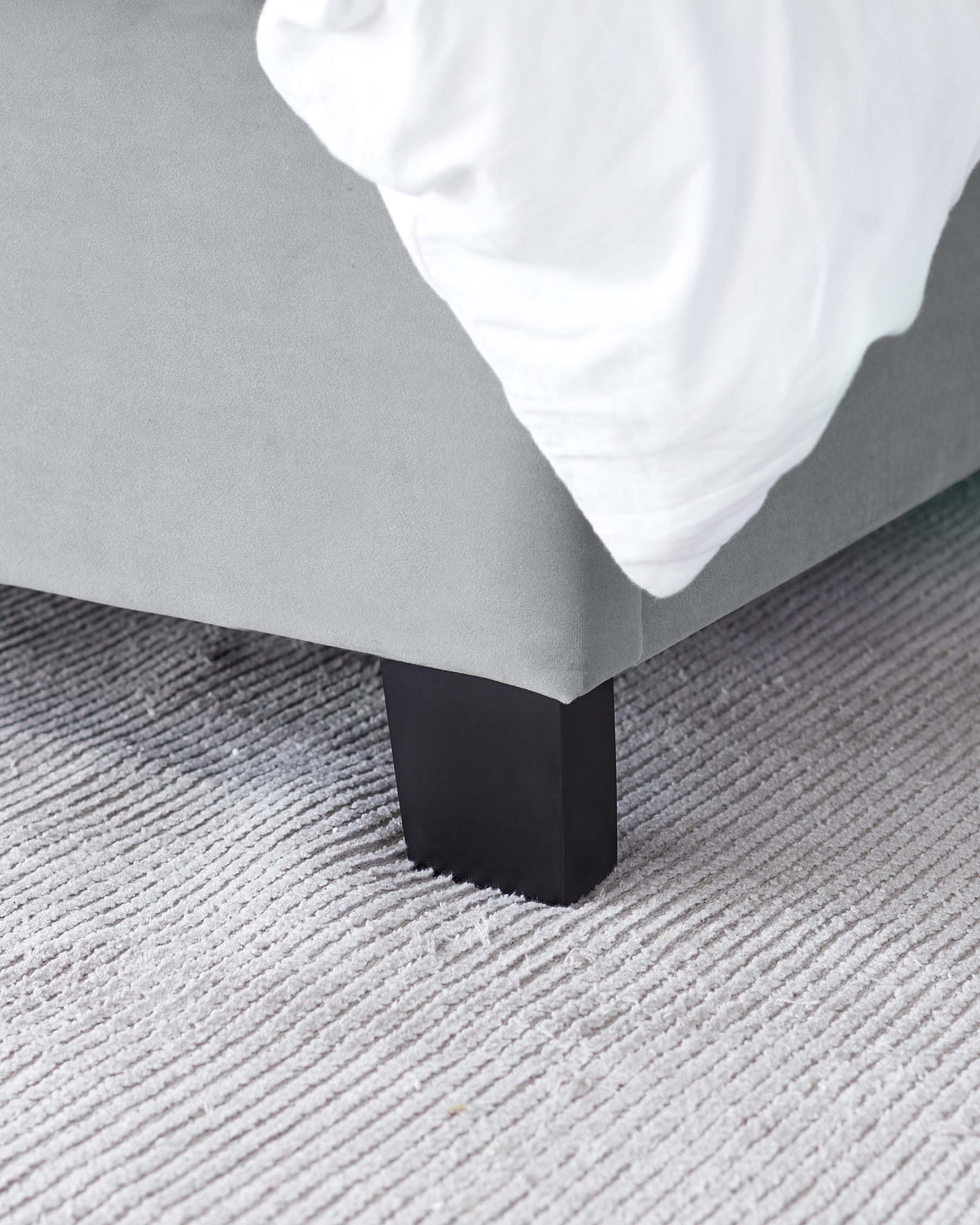 Modern minimalist style bed frame with a grey upholstered base and a single prominent black wooden leg visible, set against a textured grey carpet. White bedding drapes over the edge of the bed.