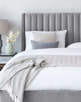 Elegant grey upholstered bed with a tall, channel-tufted headboard, showcasing crisp white bedding accented with a textured throw pillow and a patterned grey and white throw blanket, paired with a matching grey bedside table and a pastel green ceramic lamp.