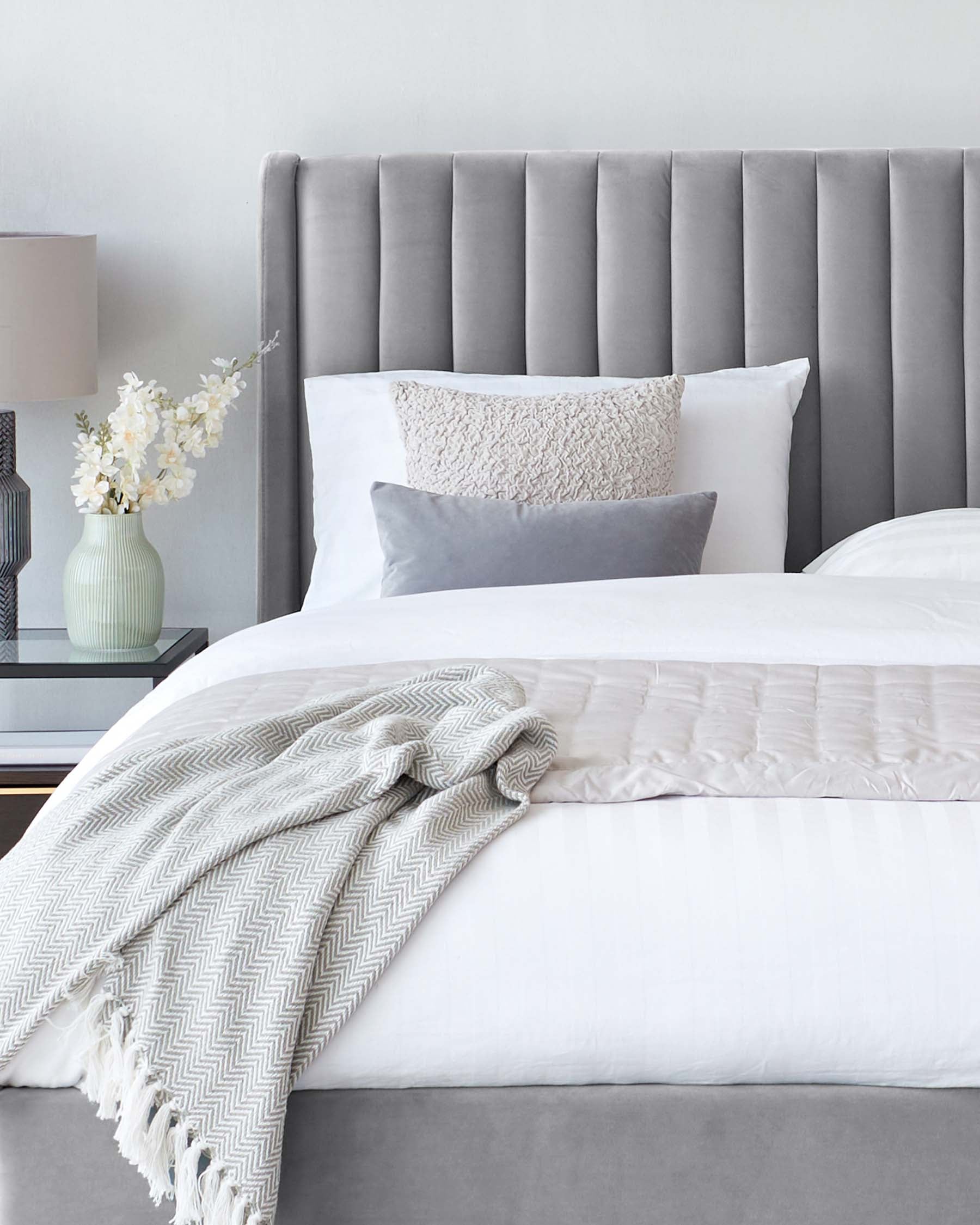 Elegant grey upholstered headboard with vertical channel tufting, white bedding with a textured throw blanket and decorative pillows, flanked by a ceramic table lamp on a nightstand.