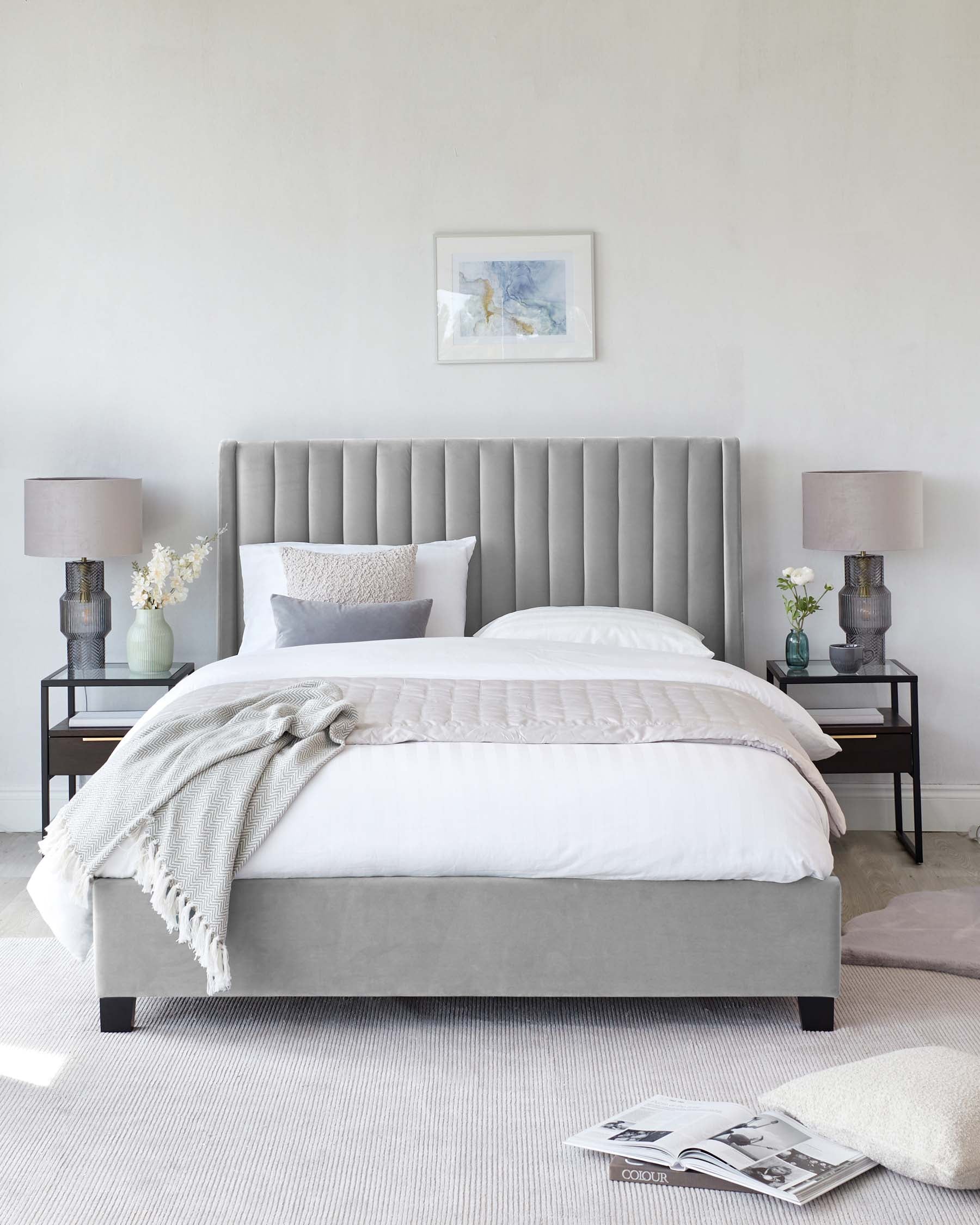 Elegant bedroom showcasing a modern, grey upholstered bed frame with a high, vertical channelled headboard, two black metal side tables with lower shelves and glass tabletops, complemented by matching grey ceramic table lamps, all set on a textured ivory area rug.