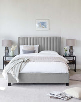 Elegant bedroom furniture set featuring a grey upholstered bed with a tall, vertical channel-tufted headboard and two black metal nightstands with glass tops, each adorned with a textured lamp and floral arrangements. A white duvet with pillows and a knit throw blanket completes the serene setting.