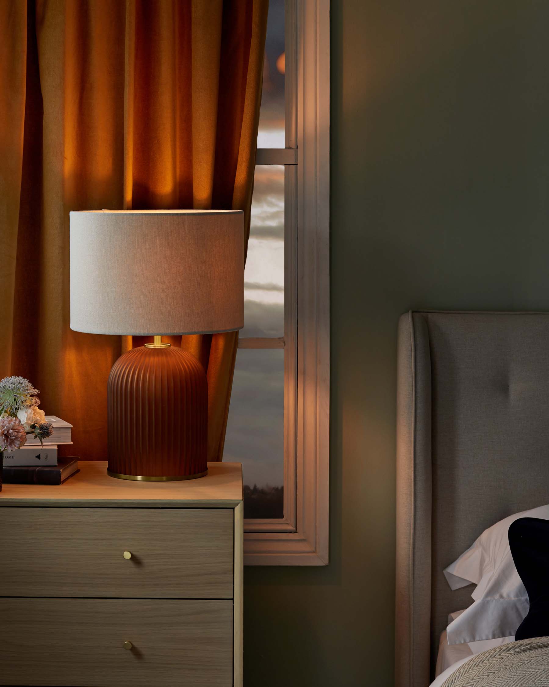 Elegant bedroom scene featuring a modern wood bedside table with a smooth finish and metal handles, topped with an orange-brown ribbed ceramic table lamp with a beige shade. There is a glimpse of a bed with grey upholstered headboard on the right.