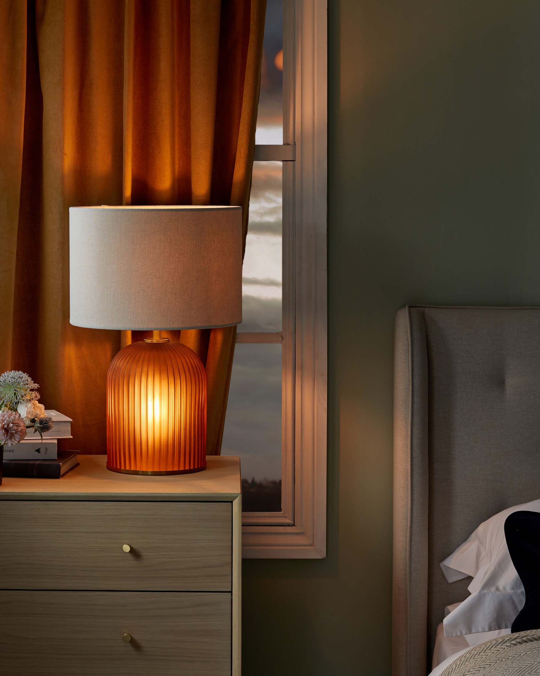 A modern bedside setup featuring an elegant wooden nightstand with two drawers, showcasing round brass knobs, and topped with a stylish, warm-glow table lamp with a grooved, orange base and cream cylindrical shade. The scene is completed with a hint of an upholstered grey headboard, suggesting a contemporary and cosy bedroom atmosphere.