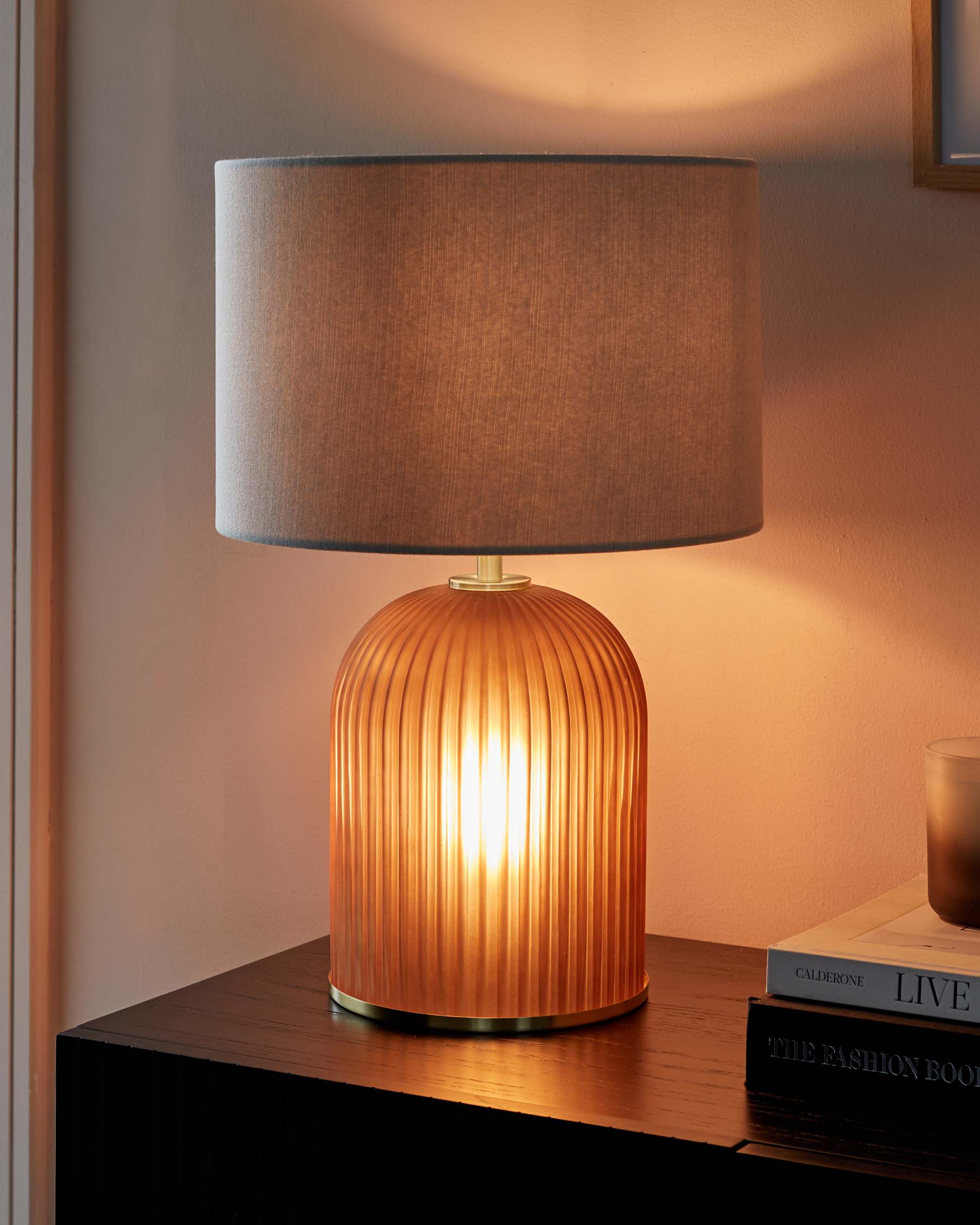 Modern table lamp with a ribbed, amber glass base and a beige fabric drum shade, placed on a dark wooden side table.