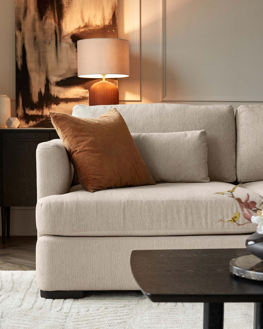 Modern living room featuring a neutral-coloured fabric sofa with plush cushions and a soft brown throw pillow. In the background, there is a dark wooden side table with a ribbed, terracotta-coloured base supporting a lamp with a beige shade. The scene includes a front view of a contemporary, dark wooden coffee table with a rounded triangular shape. The setting is completed with a textured off-white area rug. The decor creates a warm and inviting atmosphere.