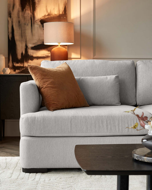 A modern minimalist living room featuring a light grey fabric sofa with clean lines and a plush look, adorned with a single rust-coloured throw pillow. A contemporary black oval coffee table sits in the foreground, while behind the sofa, a sideboard is partially visible, topped with an elegant ribbed base table lamp with a large, neutral-coloured lampshade. The scene is softened by a textured off-white area rug underfoot.