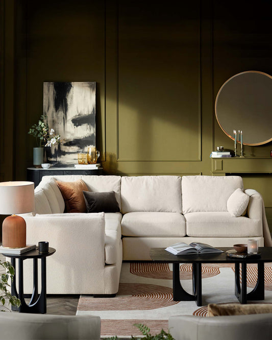 A contemporary living room featuring a plush L-shaped cream sectional sofa with assorted throw pillows, flanked by a round terracotta-coloured table lamp on a small black side table on one end. In the centre lies a pair of nesting coffee tables in black with a contrasting wood finish on top, accompanied by books, a small bowl, and a lit candle. The scene is set against a backdrop of olive green walls, with an abstract monochrome painting and a round mirror as decorative accents.