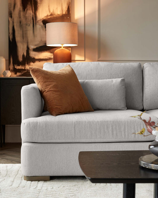 Contemporary light grey fabric sofa with clean lines and cushioned backrest, adorned with a brown decorative pillow. Beside the couch is a dark wooden side table with a ribbed, cylindrical base and a beige lampshade atop it, complementing an abstract art piece on the wall. In the foreground, partially visible is a matte black coffee table with a rounded rectangular top. The scene is set on a textured off-white rug, creating a warm and modern living space.