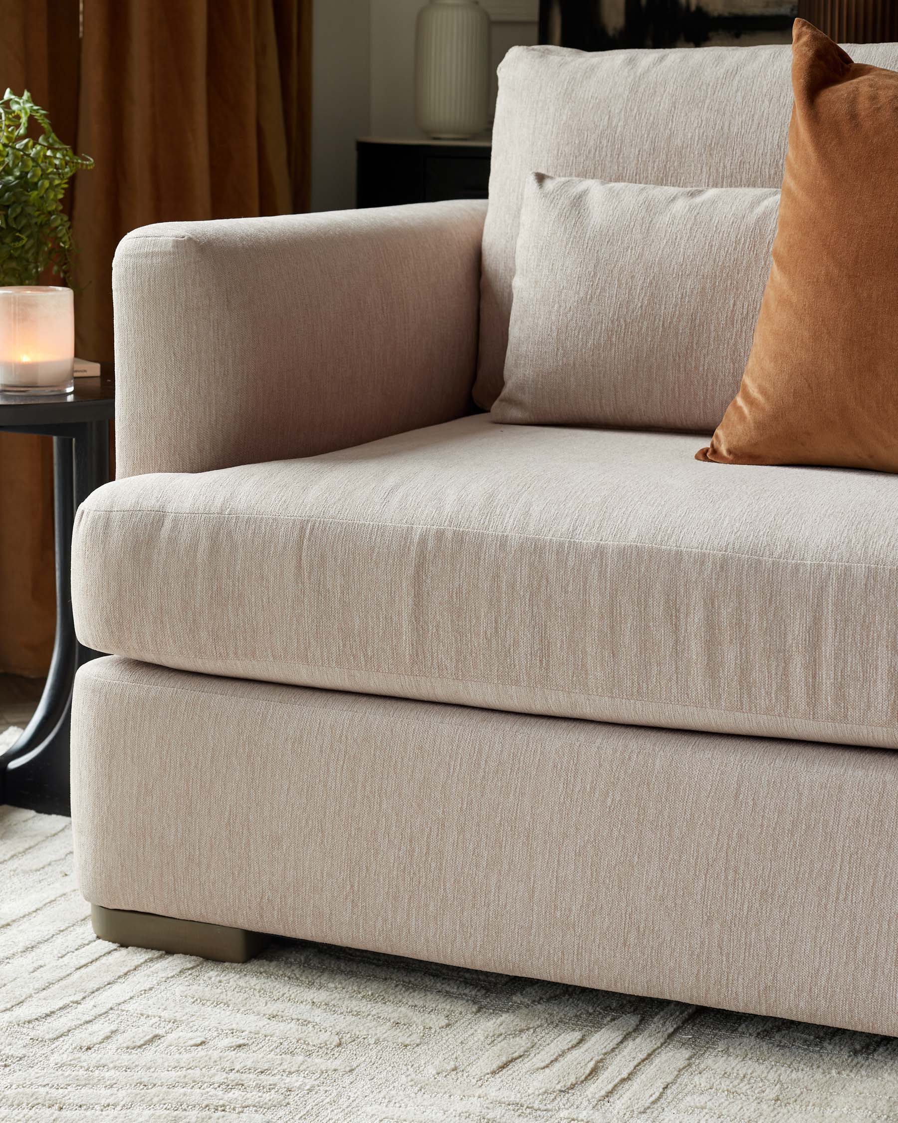 Contemporary beige fabric sofa with a clean-lined silhouette, featuring a single oversized cushion, textured upholstery, and low-profile square feet, complemented by a burnt orange velvet throw pillow. A small round black side table with a vase and lit candle is visible to the left, against a backdrop of a dark-toned cabinet and mustard drapes. The setting includes a plush cream-colored rug underfoot.