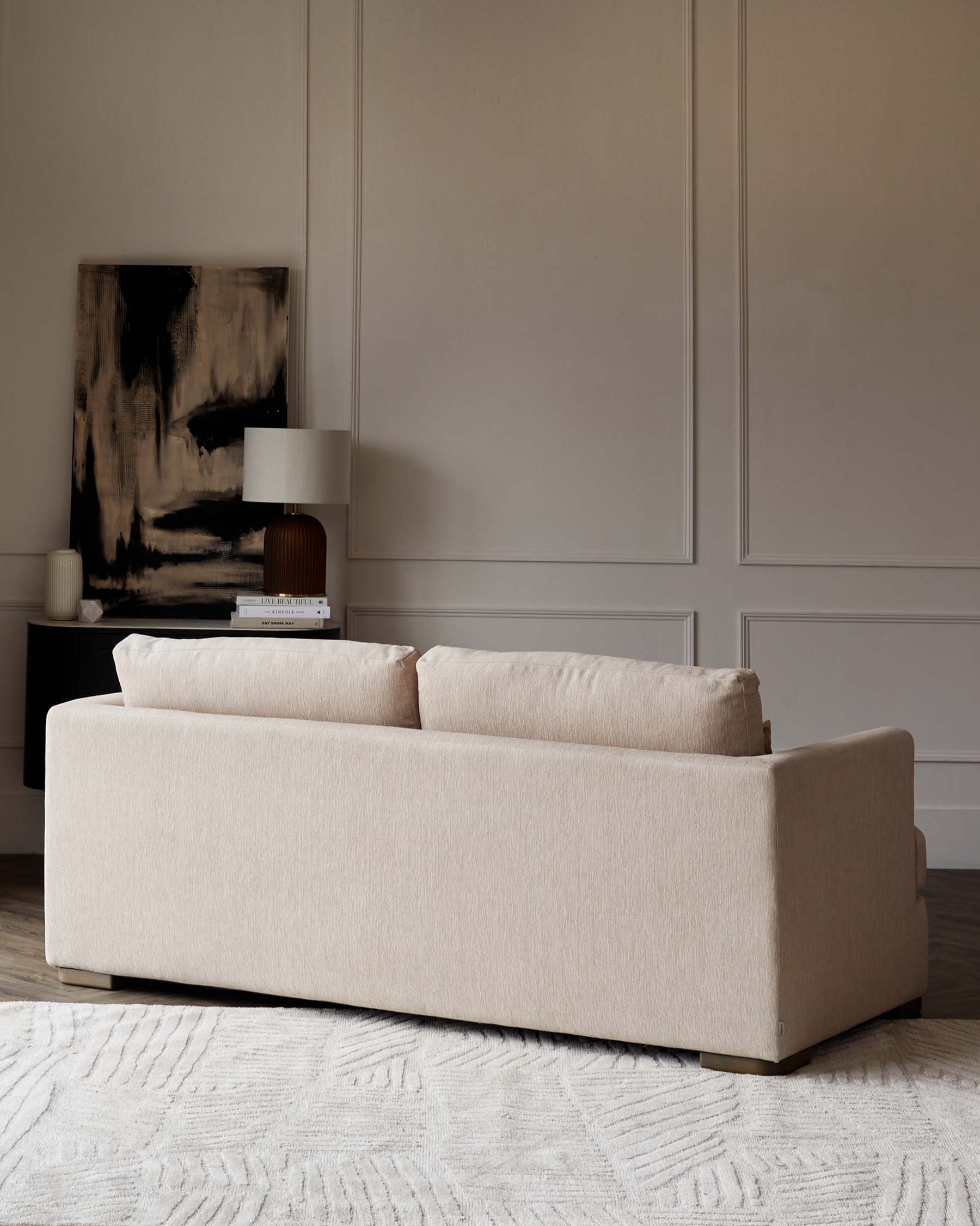 Modern neutral-toned fabric sofa with clean lines and minimalistic design, resting on a textured off-white area rug, accompanied by a contemporary dark wood side table featuring a unique lamp with a cylindrical shade, set against an elegant beige wall complemented by a large abstract monochromatic artwork.