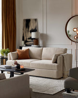 Elegant living room featuring a beige contemporary three-seater sofa with plush cushions and an amber throw pillow, complemented by a soft beige accent chair in the foreground. A dark wood oval coffee table sits at the centre atop a textured white area rug. Additional accents include a round wall mirror with a unique circular light fixture, a modern abstract painting, and a sleek black side table bearing a white ceramic lamp.