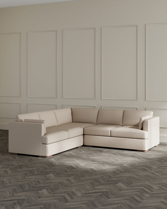 Modern beige L-shaped sectional sofa with clean lines and plush cushions, set against a panelled beige wall on a dark herringbone-patterned floor.