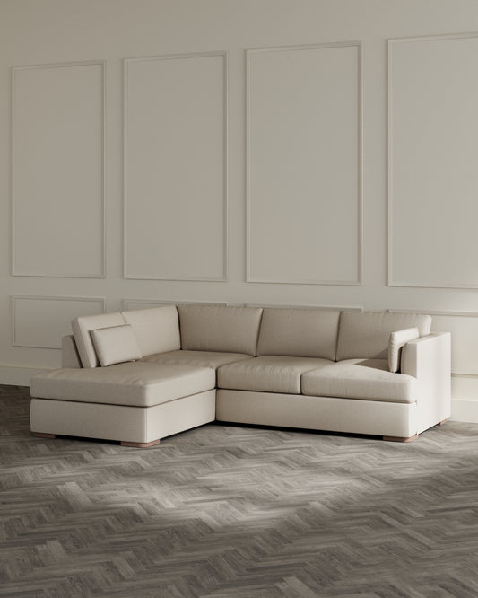 Contemporary L-shaped sectional sofa with chaise in a neutral beige fabric, featuring clean lines, plush cushions, and a minimalist design with low, squared armrests and subtle base support in a contrasting wood finish.