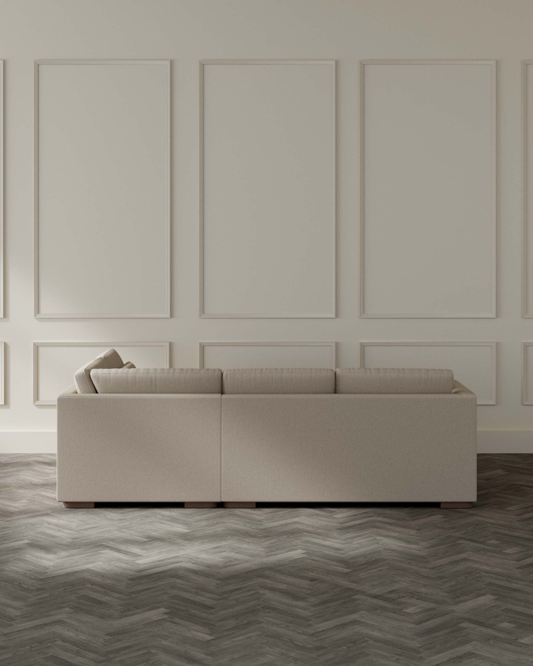 Contemporary beige upholstered modular sofa with clean lines and minimalistic design, featuring plush back cushions and structured seat cushions, resting on low-profile brown wooden legs in a spacious room with elegant panelled walls and herringbone wood flooring.