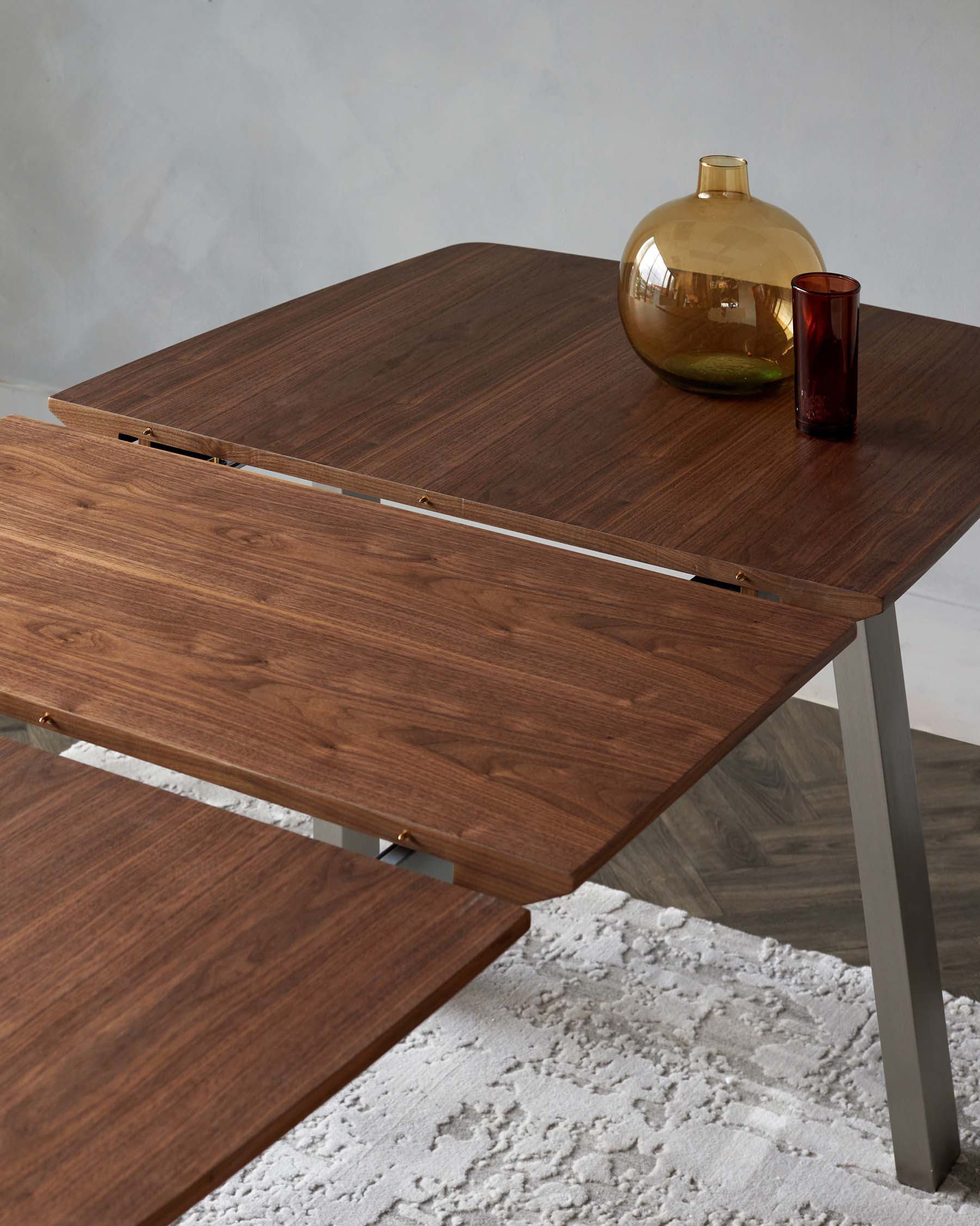 A contemporary extendable dining table with a dark walnut wood finish and metallic accents on the expansion mechanism. The table is partially extended, displaying its unique design feature. A large gold-tinted glass vase and a smaller red glass sit atop, adding a touch of colour to the elegant setup. The table is set on a textured grey rug, contrasting with a neutral wall in the background.