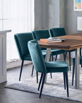 A modern dining set featuring a rectangular walnut table and six plush teal upholstered dining chairs with vertical channel stitching and tapered black legs. The set is positioned atop a textured light grey area rug, and the table is adorned with a stack of magazines and books, presenting an elegant and contemporary dining arrangement.