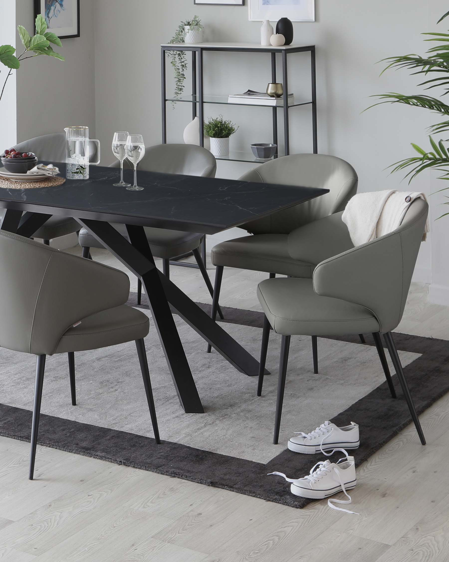 Modern dining room with a rectangular black marble table and four taupe upholstered chairs with black metal legs, set on a dark grey area rug. In the background, a minimalist metal framed bookshelf displays decorative items.