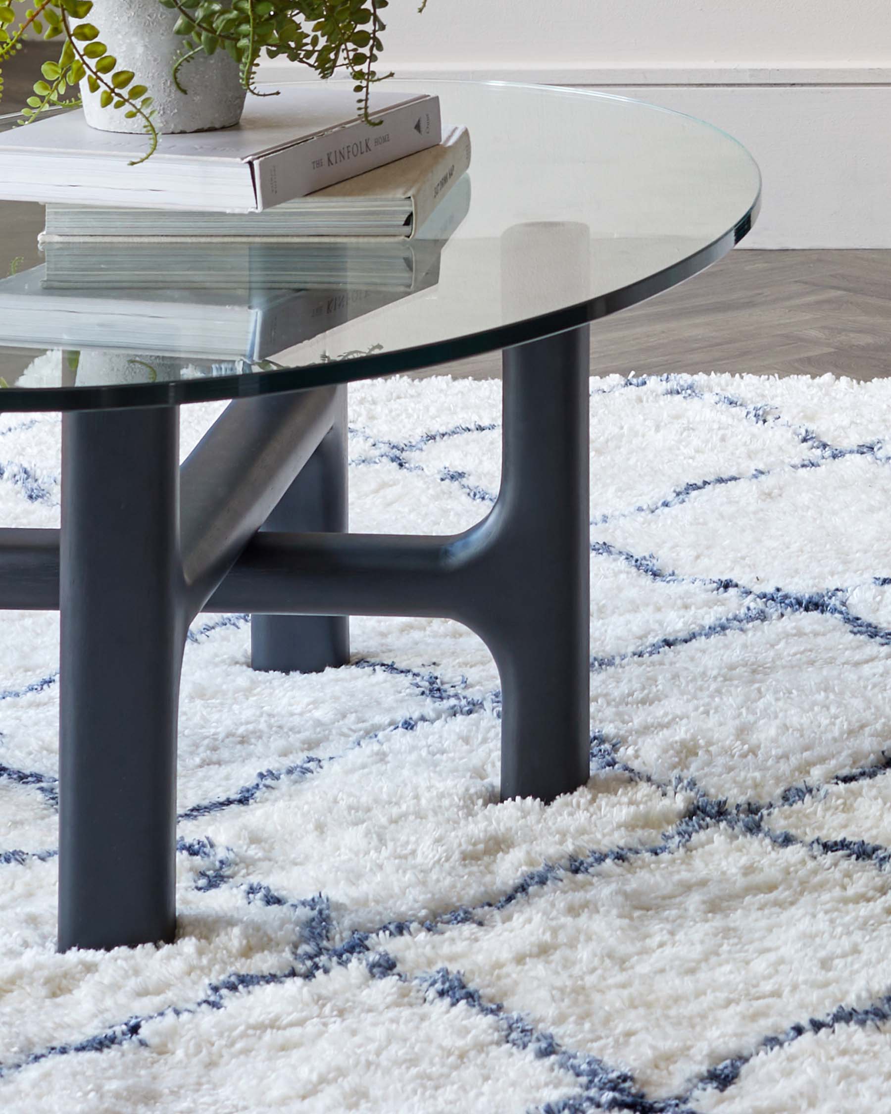 Modern round glass-top coffee table with a unique, matte black metal base structure, set on a textured white rug with blue accents.