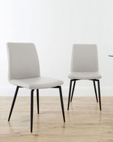 Two minimalist gray dining chairs with sleek black legs, set against a light background and wooden floor.