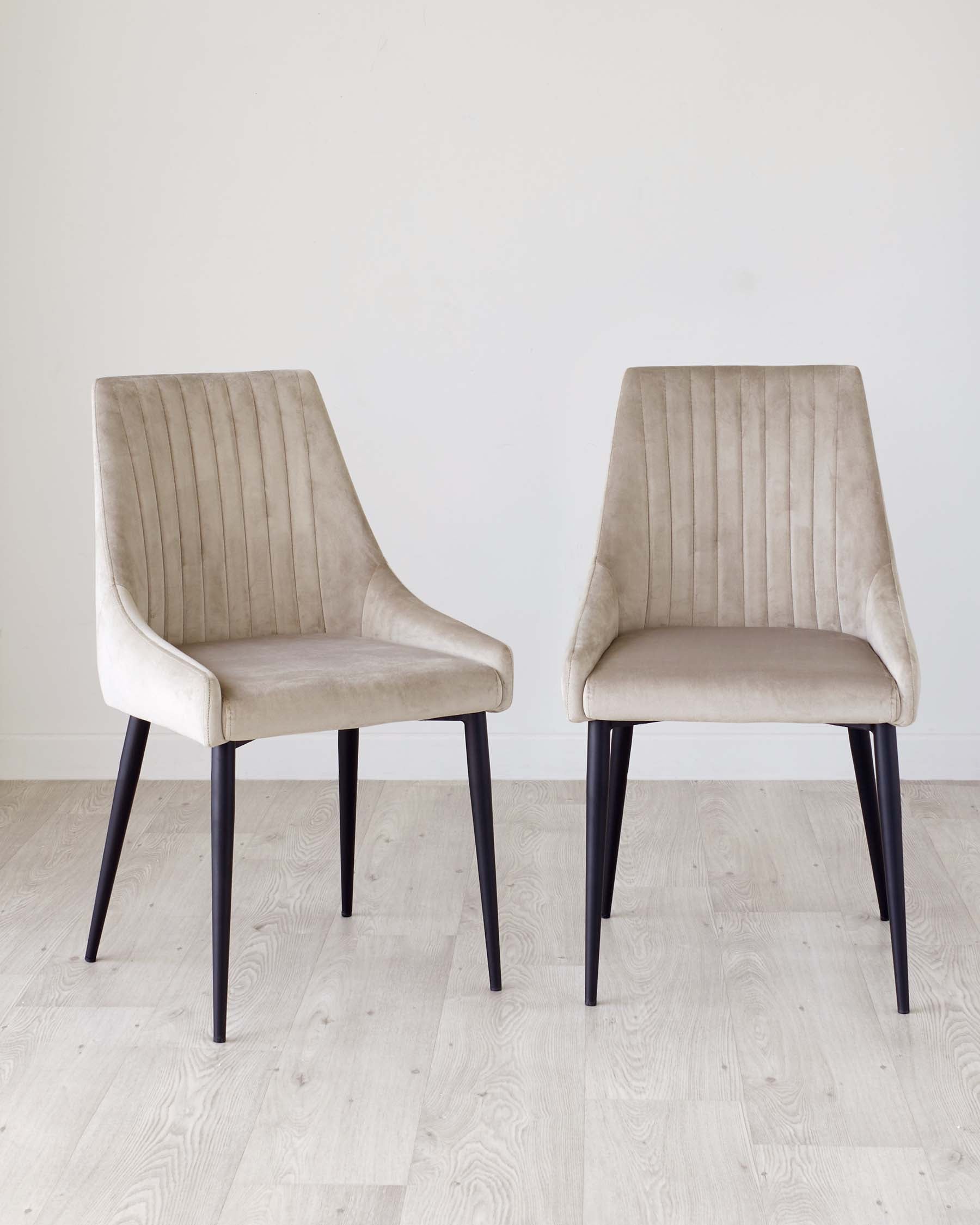 Two modern beige upholstered chairs with vertical stitching and black legs, displayed on a light wood floor against a neutral wall.