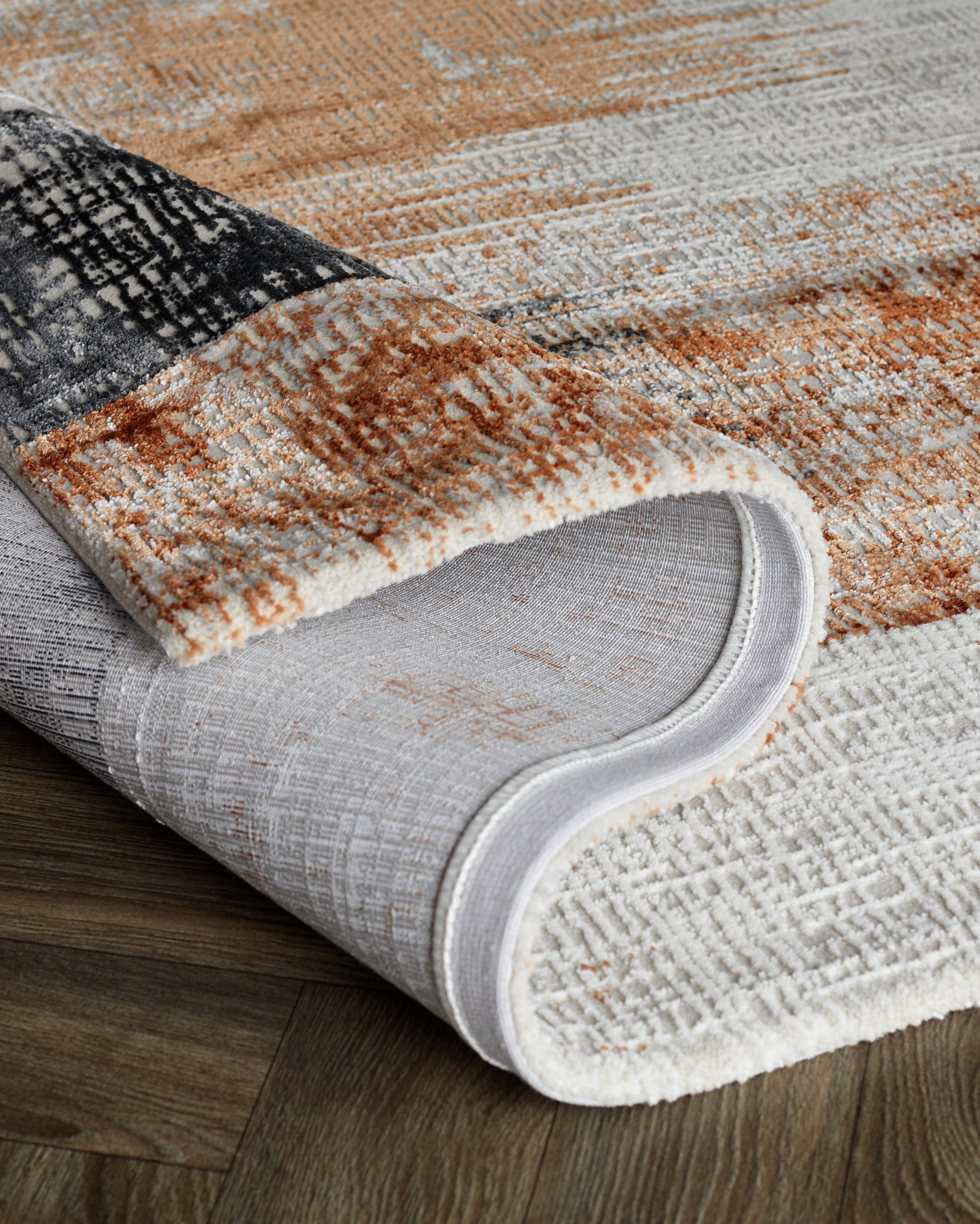 Textured area rugs with a combination of neutral tones; featuring patterns of grey, black, white, and shades of tan and brown on a wooden floor. One rug is rolled partially to show layers and thickness.