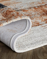 A rolled corner of a textured area rug with a pattern in shades of burnt orange, grey, and cream, lying on a dark wood floor.