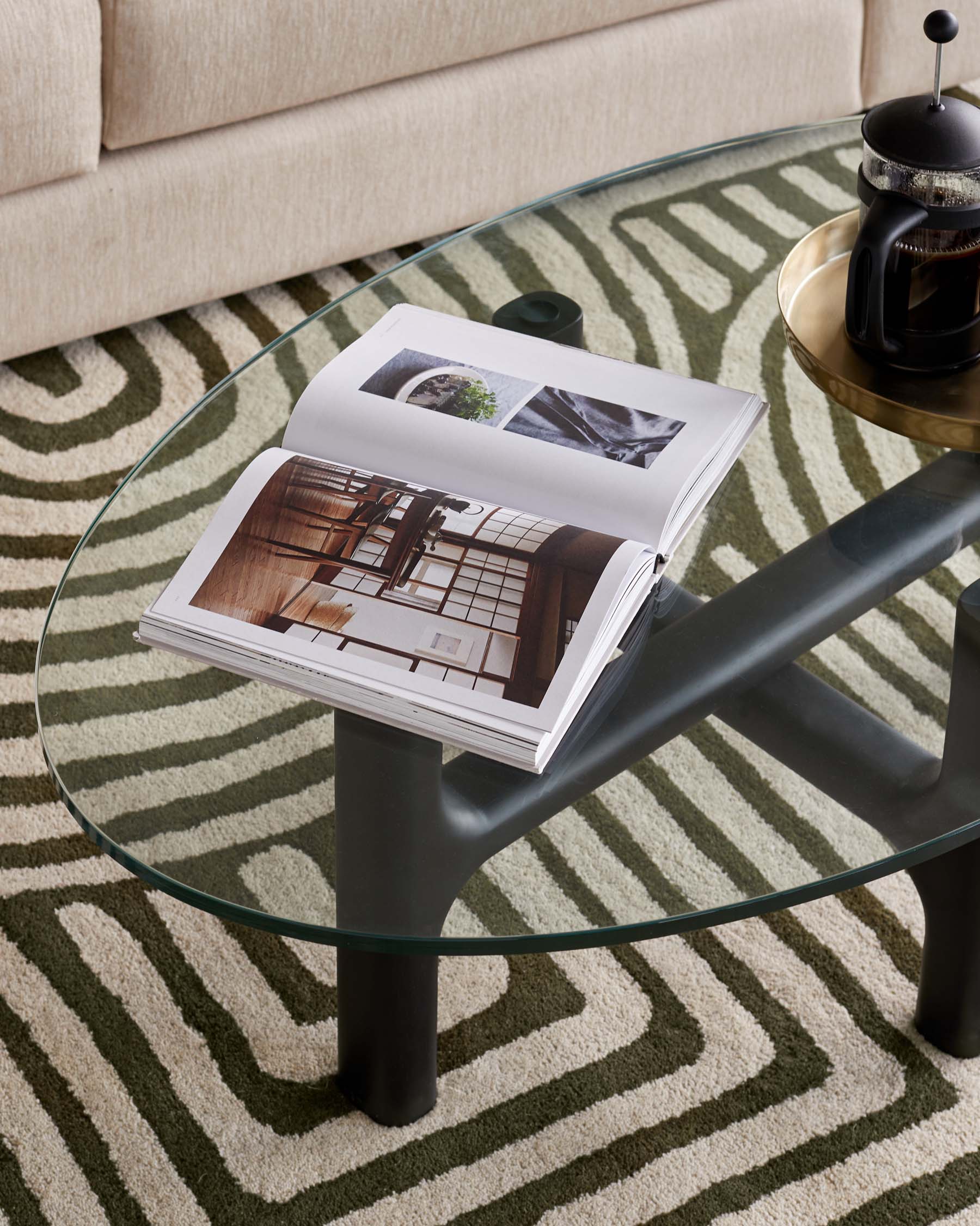 Round glass-top coffee table with three cylindrical black legs on a patterned beige and green carpet, near an off-white sofa, adorned with a large open book and a French press on a brass tray.