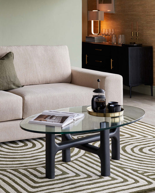 Modern living room with a minimalist beige sofa, round glass top coffee table with unique dark wood base, and a sleek black sideboard with brass handles.