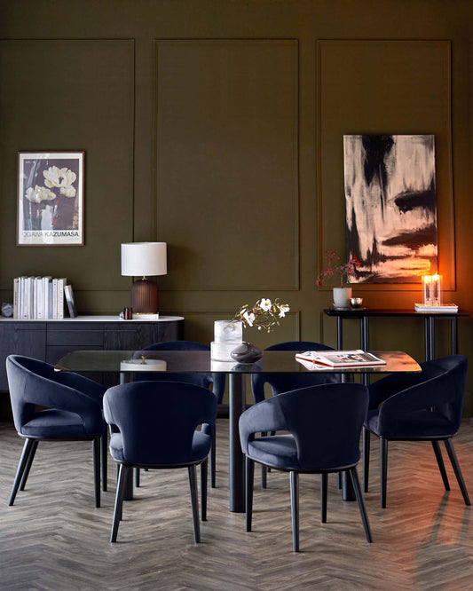 A modern dining setup featuring a glass table and four navy upholstered chairs, accented by a decorative lamp and artwork.