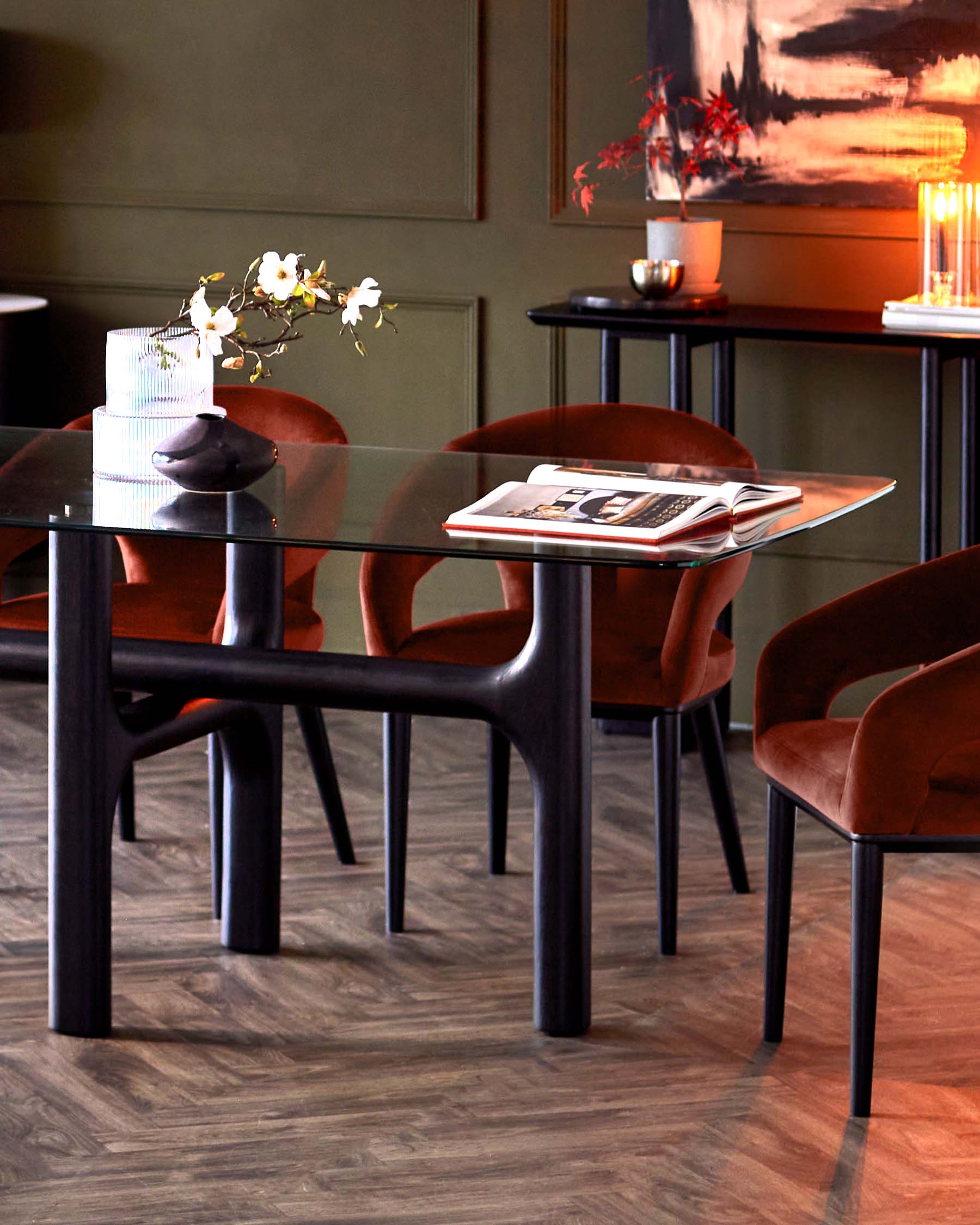 Stylish glass-top dining table with dark wood legs, complemented by plush rust-colored chairs and elegant decor accents.