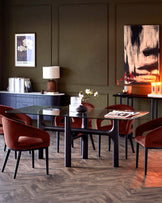 Glass-top dining table with sleek black legs and plush burgundy chairs, complemented by decorative pieces and a dark green wall.