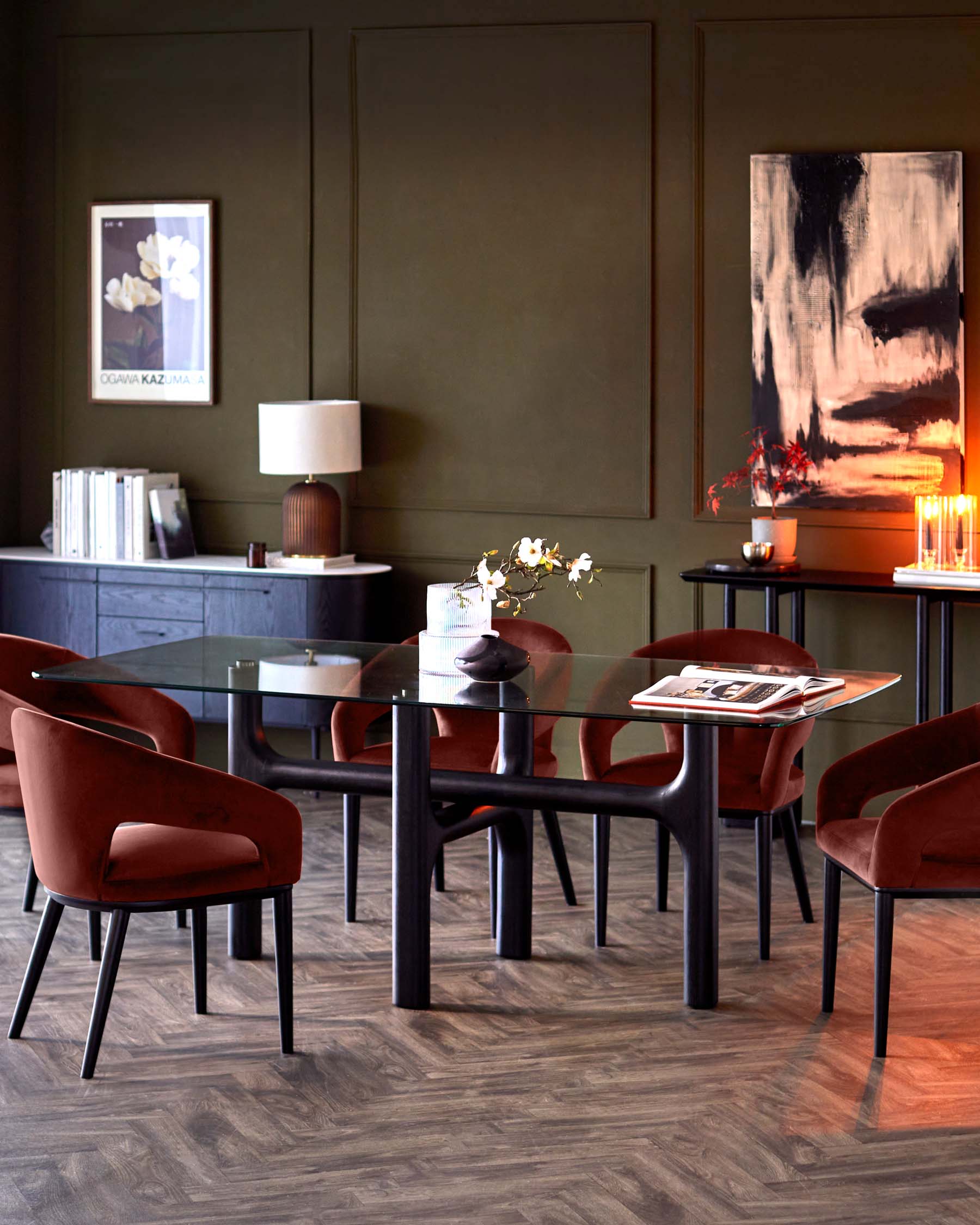 Glass-top dining table with sleek black legs and plush burgundy chairs, complemented by decorative pieces and a dark green wall.