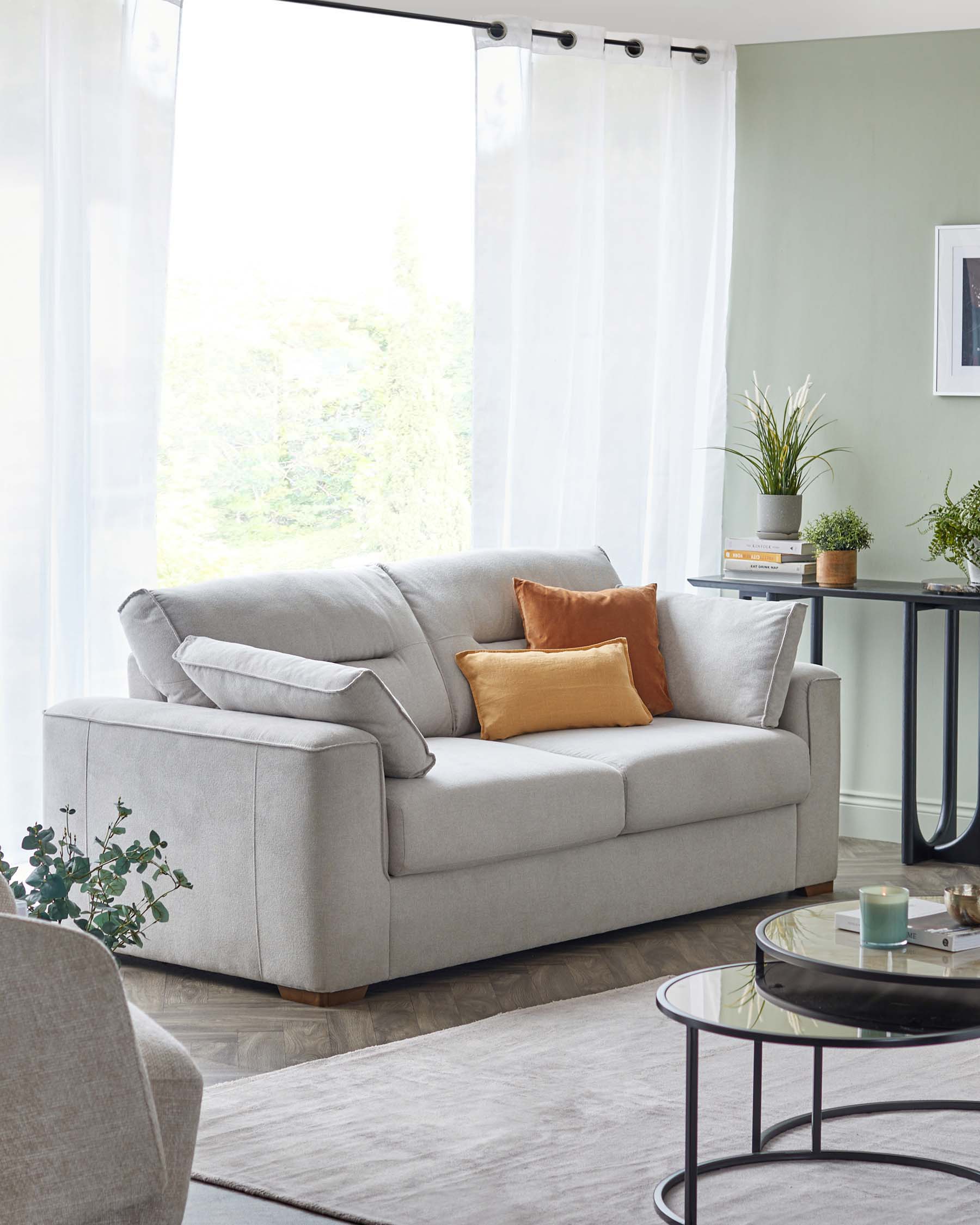 Contemporary-style two-seater sofa with plush light grey upholstery and tapered wooden legs. cosy throw pillows in white and mustard complement the sofa, which is adjacent to a sleek round side table with a dark glass top and black frame. A round glass-top coffee table with a black metal frame sits on a neutral-toned area rug, enhancing the modern and inviting ambiance of the room.