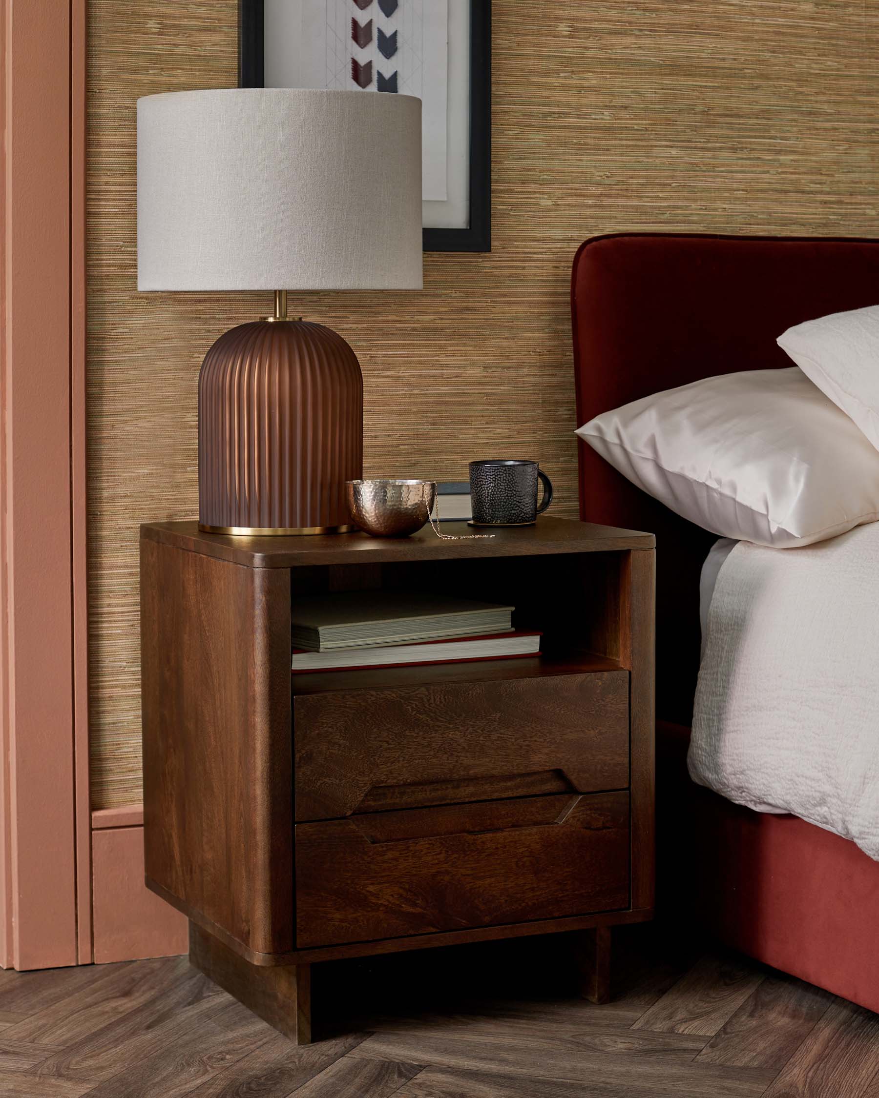 A modern bedside table in a rich wood finish, featuring two drawers and a stylish lamp with a beige shade and ribbed base.