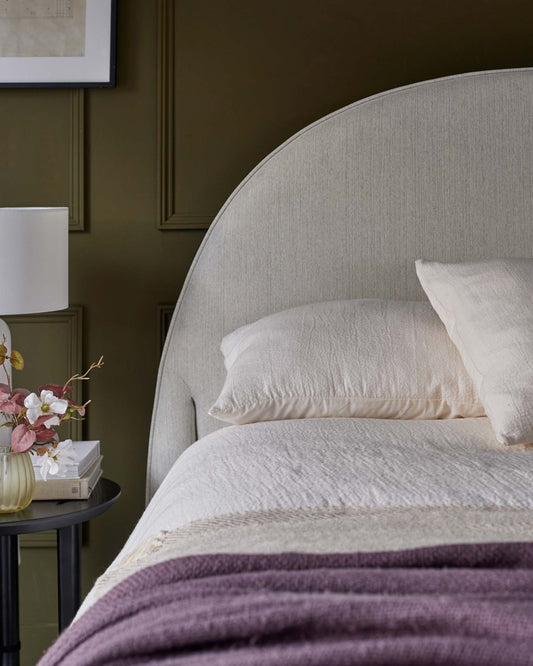 Elegant bed with a curved, light grey upholstered headboard, soft bedding, and a round side table featuring a floral arrangement.