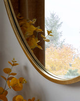 Elegant wall mirror with a reflective golden frame gently curving into an oval shape, set against a light-coloured wall and reflecting autumnal trees visible through a nearby window.