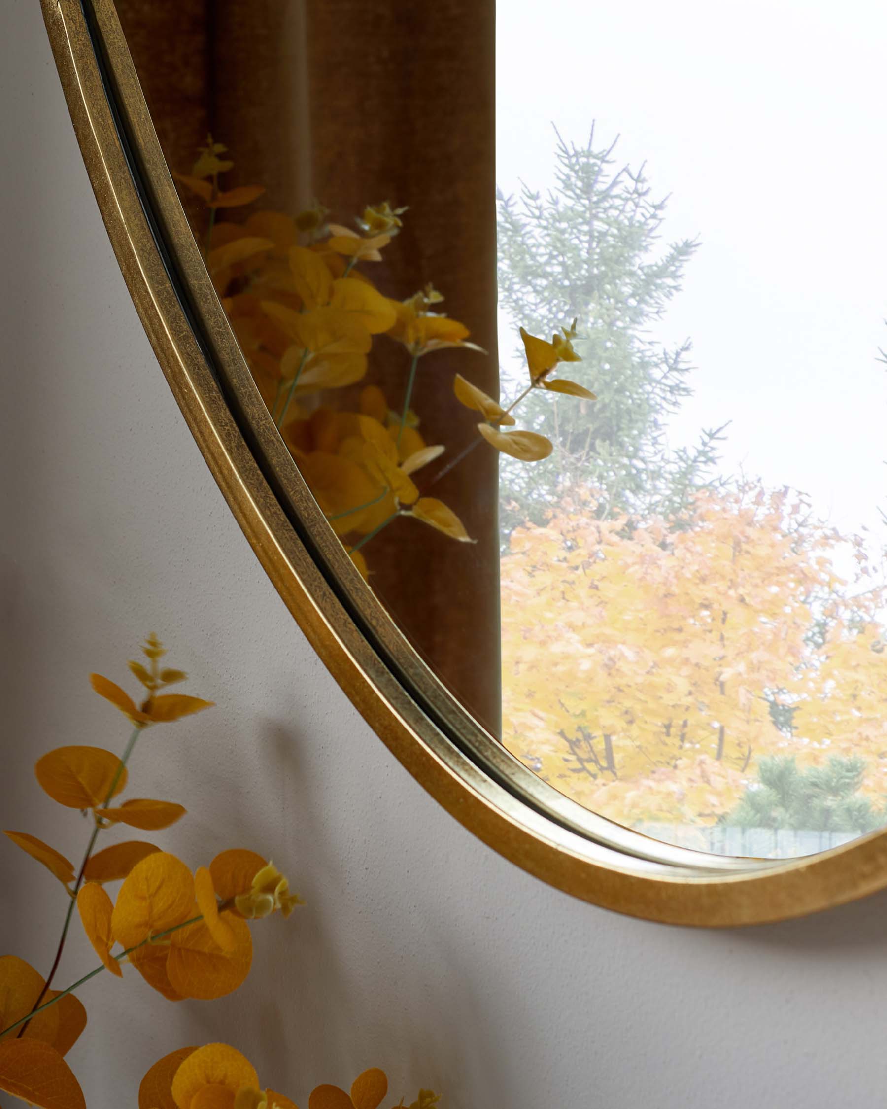 Elegant wall mirror with a reflective golden frame gently curving into an oval shape, set against a light-coloured wall and reflecting autumnal trees visible through a nearby window.