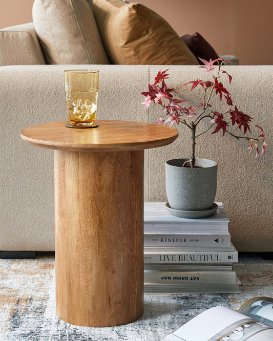 A cylindrical wooden side table holds a glass, beside a plant in a speckled pot and stacked books, with a cozy sofa in background.