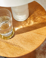 A round wooden side table with a smooth finish, featuring a glass of water and a white lamp atop it.