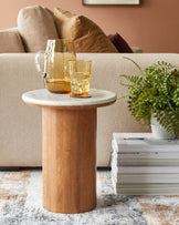 Round side table with a marble top and wooden base, complemented by a pitcher and glass, alongside stacked magazines and a plant.