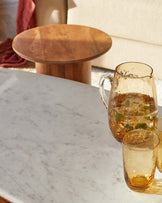 A round wooden side table, a marble topped coffee table, a glass pitcher, and matching amber-hued drinking glasses.