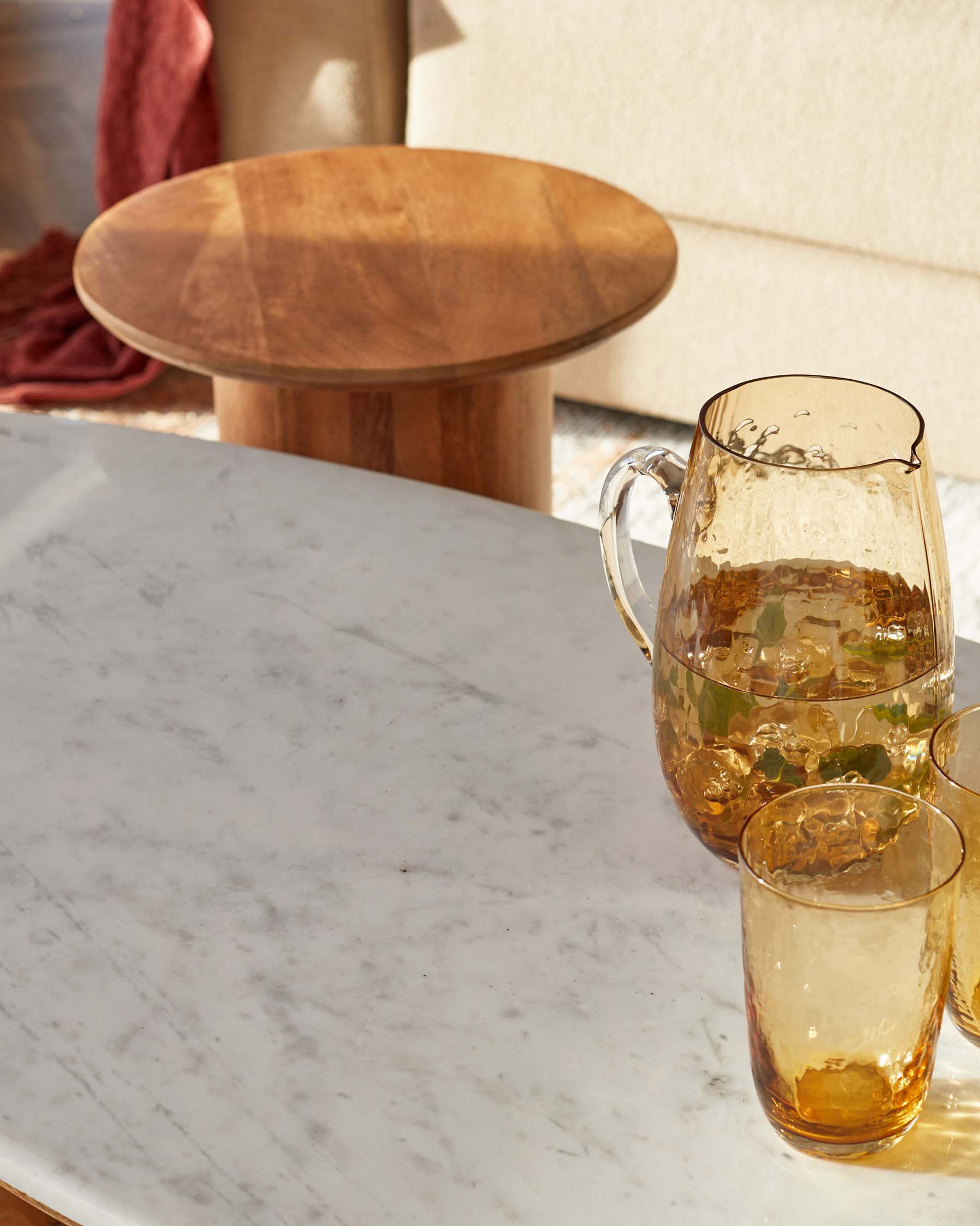 A round wooden side table, a marble topped coffee table, a glass pitcher, and matching amber-hued drinking glasses.