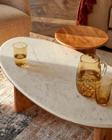 A sleek marble coffee table with wooden legs, paired with a round wooden side table, both accented by amber glassware.