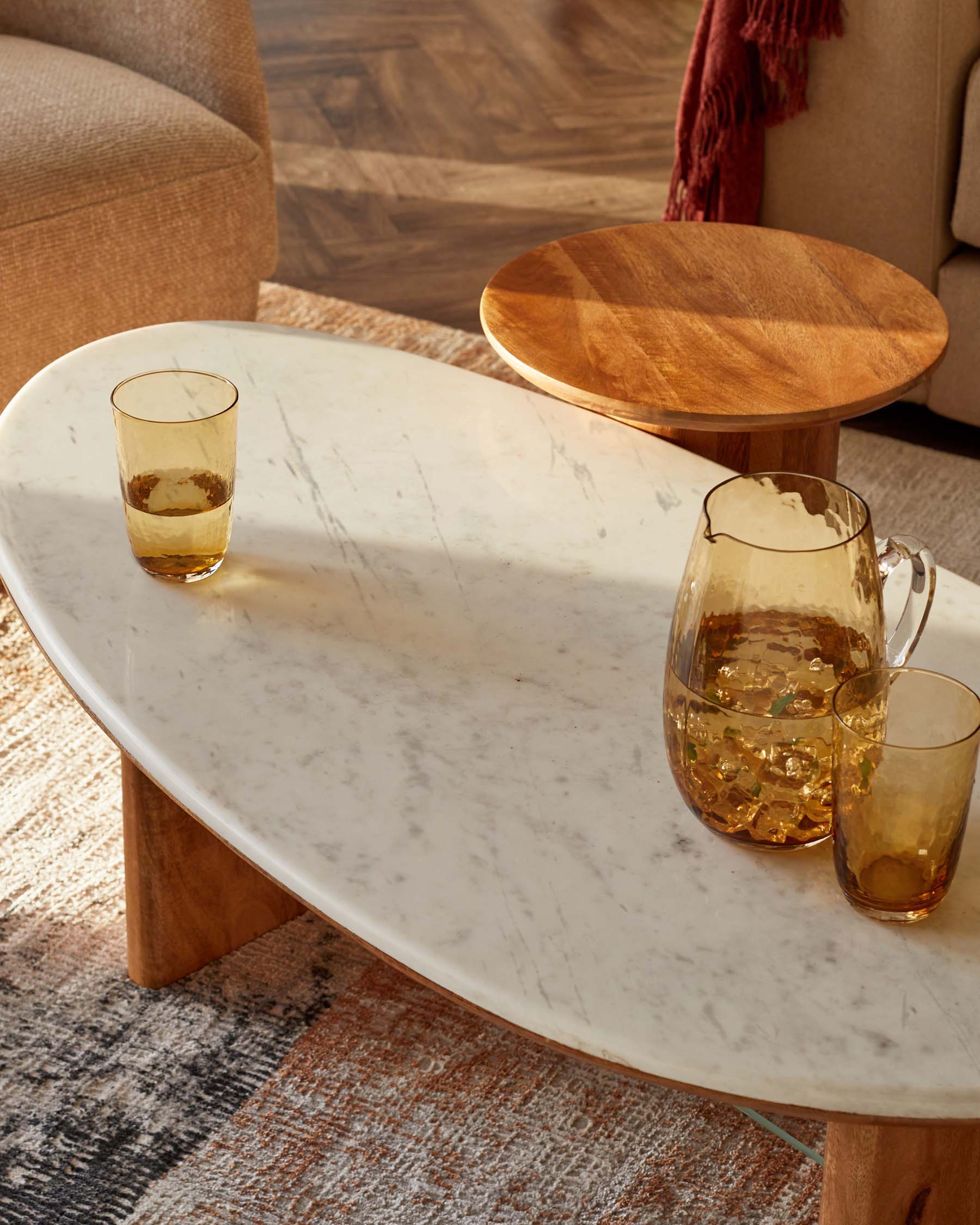 A marble-top coffee table with wooden legs, paired with a round wooden side table, featuring amber glassware.