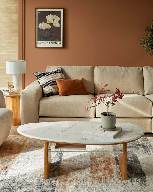 Cozy beige sofa with orange and patterned cushions, a marble coffee table with wood legs, and a decorative plant.